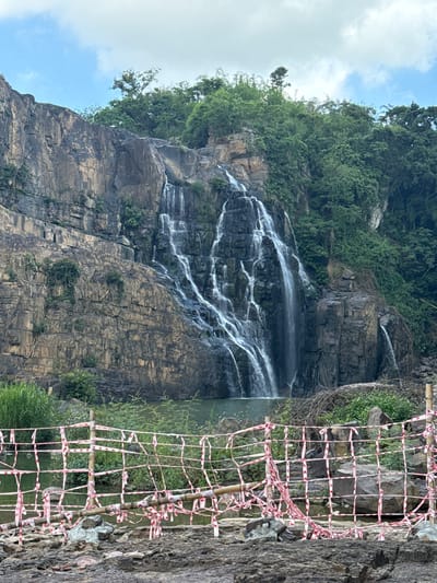 The image depicts a majestic waterfall cascading down a rugged, rocky cliff face. Lush green vegetation covers the top of the cliff and clings to its sides in places. In the foreground, a rustic barrier constructed from bamboo poles and red and white striped tape cordons off the area, suggesting it's a viewing point or a restricted zone. The water collects in a pool at the base of the waterfall. The sky above is partly cloudy with patches of blue. Based on the context provided, this scene is located in the city of Tân Phú, Vietnam, likely within a natural park or recreational area known for its waterfalls.
