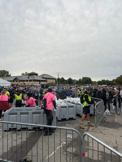 The image depicts an outdoor event scene located in a paved area adjacent to the Weymouth College campus in Weymouth, United Kingdom. The Weymouth College buildings, characterized by their multi-story structure, dark gabled roofs, and light-colored stone or brick facades, are visible in the mid-background beneath an overcast, grey sky.
In the foreground and mid-ground, a large gathering of individuals is observed. Numerous bicycles are racked in dense rows, suggesting a transition area for a cycling event or triathlon. Several grey, rectangular plastic containers are prominent, with multiple units visible in a row on the right and at least one on the left. These containers hold numerous white, uniformly sized bags, likely participant gear.
Multiple individuals are actively engaged:
- A man wearing a bright pink t-shirt is leaning into one of the grey containers on the left, appearing to interact with its contents.
- Another man in a bright pink t-shirt with a black backpack is standing with his back to the camera, next to the containers on the right.
- A man in a yellow high-visibility vest labeled "TEAM" is visible on the far left.
- A man in a yellow high-visibility vest and tan boots is standing on the right, holding onto a metal crowd control barrier.
- Other individuals in the crowd are wearing a mix of casual attire and athletic wear, with some sporting bib numbers.
Metal crowd control barriers, some branded "e.g. Events," delineate sections of the paved area in the immediate foreground and mid-ground. The ground is a dark, paved surface, likely asphalt, with some faint white markings. Trees and other vegetation are visible behind the college buildings.
