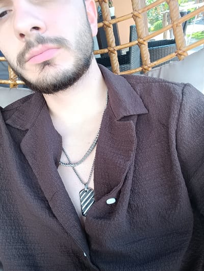 A close-up shot depicts a person wearing a textured, dark brown shirt or jacket. The garment is partially buttoned, revealing a silver necklace with a pendant. The person has a beard and is positioned indoors or in a covered outdoor area, with a wicker chair or structure visible in the background. The location is not discernible from the image alone.