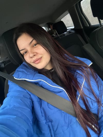 A young adult female is depicted in a close-up, chest-up shot from within a vehicle. She has fair skin, dark brown long hair parted on the left, and visible freckles across her nose and cheeks. Her expression is a slight smile directed towards the viewer. She is wearing a bright blue puffer jacket with white zipper details over a black garment, and a black vehicle seatbelt is positioned diagonally across her chest. The interior of the vehicle shows dark fabric seats, including a headrest behind her and one further back on the passenger side. A blurred figure is partially visible in the rear seat directly behind the front passenger seat. The exterior environment through the vehicle windows is indistinct. The precise location within the city of Padarevo, Bulgaria, cannot be determined from the visual information provided.