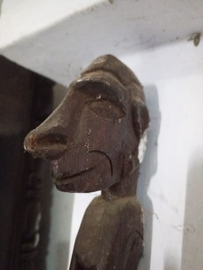 The image displays a dark brown wooden carving, viewed from an oblique angle, located indoors in Keo Kouang, Laos. The carving represents the head and upper torso of a stylized figure, oriented in profile facing left. Distinct features include an elongated, prominent nose, recessed ovoid shapes for eyes, and an incised curved line defining the mouth and chin area. The top of the head exhibits multiple parallel horizontal grooves. The wood surface presents a dusty patina, with scattered white particulate matter and localized areas of white discoloration, notably on the right side of the head and the nasal bridge. Fine cobweb strands adhere to the carving and the adjacent surfaces. The background consists of a predominantly white, slightly textured vertical plane to the right and above the carving, and a darker, indistinct vertical element to the left. The scene is illuminated from the viewer's upper right, producing a soft shadow of the carving on the white background.