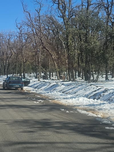 The image displays a segment of a two-lane asphalt road in Ait Naamane, Morocco, bordered by a snow-covered forest under a clear blue sky. A dark blue four-door sedan is parked on the left shoulder of the road, oriented away from the viewer, with its front driver's side door open. Substantial snow banks, discolored with dirt in sections, line the right side of the road and extend into a dense forest. The forest consists of predominantly bare deciduous trees and a lesser number of evergreen trees with dark foliage. Long, dark shadows from the trees are cast across the road surface. No individuals are present in the depicted scene.