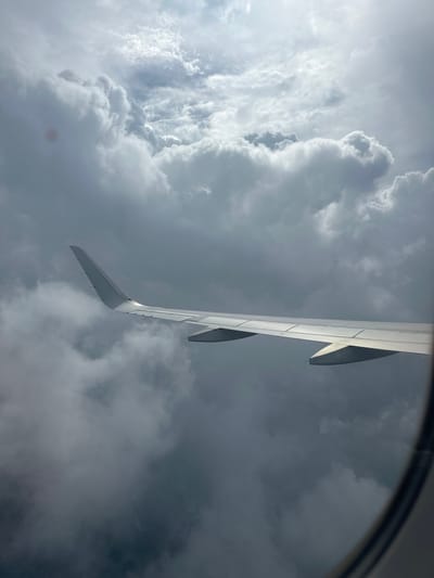 The image shows an airplane wing and turbulent clouds from a window. The scene is from an airplane in flight over Fogang County, China. The clouds are gray and dense, with some patches of lighter sky visible. The airplane wing is white and extends across the frame. The image is taken from inside the airplane, looking out the window.