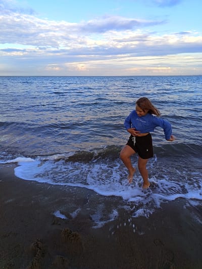 The image captures a young girl standing on a sandy beach at the edge of the sea. She is wearing a blue cropped sweatshirt and black shorts. The girl is in motion, with her left leg raised as if mid-stride, and her right arm extended. The water is a deep blue with gentle waves lapping onto the shore, creating white foam. The sky above is a soft blue with scattered white clouds, suggesting it is either early morning or late evening. The overall mood is playful and serene, with the girl appearing to be enjoying herself by the water. The location is Yarovoye, Russia.