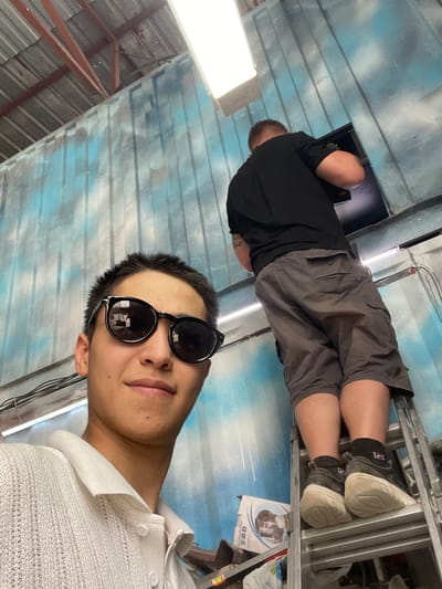 In a dimly lit interior space, potentially a workshop or storage area in Кенен Әзірбаев ауылы, Kazakhstan, two individuals are present. In the foreground, a person wearing sunglasses and a white collared shirt faces the camera, taking a selfie. Behind them, another individual, viewed from the back and standing on a metal ladder, is engaged in an activity at a high-mounted dark rectangular object. This second individual wears a black t-shirt and grey cargo shorts. The environment is characterized by corrugated metal walls painted with a blue, cloud-like pattern, and exposed ceiling beams. Electrical wires are visible along the walls, and a fluorescent light fixture illuminates the scene from above. A cardboard box with graphics is situated near the base of the ladder.