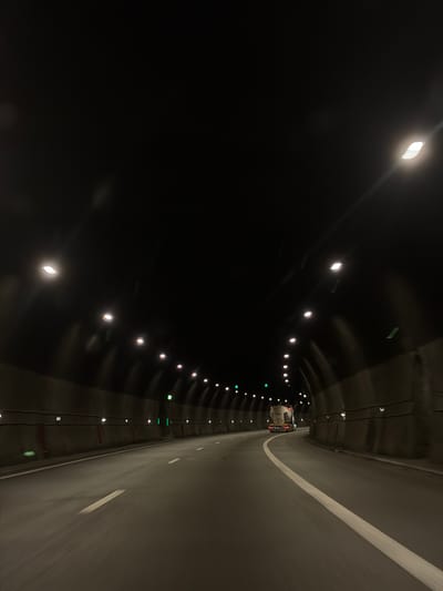 The image shows a view from inside a highway tunnel. The tunnel is dark, with artificial lights illuminating the road. The road is paved and has white lane markings. The walls of the tunnel are concrete and appear to be textured. There is a truck visible in the distance, driving in the same direction as the camera. The sky is not visible, as the image is entirely within the tunnel. This location is not in Churek, Bulgaria, as there is no information in the image to suggest this.