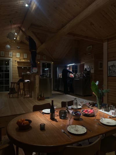 A cozy wooden cabin interior features a large oval table set for dinner, complete with plates, silverware, glasses, and fruit bowls. In the background, a person is working at the kitchen counter under bright lighting. A second person is bending down near a wall with several framed pictures and a window. The lighting is dim, creating a warm, inviting ambiance.