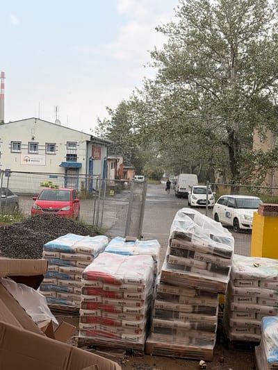 The image depicts an outdoor scene in the city of Příbram, Czechia, on a rainy day. In the foreground, stacks of bagged construction materials, likely cement or mortar, are visible. These bags are wrapped in plastic for protection. To the left, partially obscured cardboard boxes suggest recent delivery or unpacking. Behind the materials, a chain-link fence encloses a construction site. Beyond the fence, a red car is parked near a white building. This building appears to be an industrial or commercial structure with several windows and a sign that reads "MUSIC CLUB #beckco". Further down a paved road, several vehicles are present, including white vans and cars. A person is walking on the wet road. A large, mature tree with lush foliage dominates the background, with a tall industrial chimney visible in the far distance. The overall atmosphere is one of ongoing construction or renovation, with overcast skies and visible raindrops contributing to a somber, damp environment.