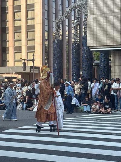 This detailed image captures a vibrant street scene in Kanazawa, Japan, showcasing a cultural procession or festival. The foreground is dominated by a black asphalt road featuring a prominent white and black zebra crossing.
Three figures in ceremonial attire are central to the scene, traversing the zebra crossing. The most prominent figure, positioned slightly right of center, is an adult wearing an elaborate, patterned orange and gold brocade robe. This individual is equipped with white tabi socks and walks on two tall, black wooden stilts, resembling elevated geta. A large, ornate golden hat or headdress crowns their head, and they hold a long, red staff capped with a distinctive silver or white blade-like ornament. Their face is partially obscured by white coloring or a mask. To the left of this central figure, another individual in a light blue traditional Japanese robe (kimono-style top and hakama-like pants) and a tall, black, flat-topped hat walks on the paved area alongside the street. A third participant, younger in appearance and dressed in a simple white robe, is positioned to the right of the stilt-walker, holding a small white stick or implement.
Numerous spectators line both sides of the street, observing the procession. They consist of a diverse group of adults and children, casually dressed in modern clothing such as t-shirts, jeans, and caps. Many are seated directly on the pavement, while others stand behind them. On the left side, a police officer in a blue uniform is visible among the crowd.
The background features urban architecture. To the left, a multi-story building with a light reddish-brown, textured facade and multiple rectangular windows, many fitted with light-colored blinds, rises. Two of these windows, on the left section of the building, contain small red triangular ornaments. To the right, a section of a building with a light-grey, horizontally tiled or bricked exterior is visible. Interspersed in the mid-ground are striking public art installations: tall, cylindrical pillars composed of dark blue, irregularly shaped mosaic tiles. From the top of these pillars, large, metallic, undulating, and stacked ribbon-like sculptures curve upwards. A black street light pole with a visible amber traffic signal is located above the crowd on the left. The scene is illuminated by bright daylight, casting distinct shadows.