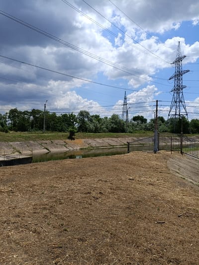 The image depicts a dry, grassy embankment next to a concrete canal filled with murky water. In the background, a line of dense green trees borders the canal. Above the trees, a bright blue sky is visible, partially obscured by large, fluffy white clouds and dramatic dark grey clouds. Several high-voltage power lines traverse the sky, supported by tall, lattice-style metal pylons. A single streetlamp on a metal pole stands near the canal's edge. A rusted metal fence runs along the top of the embankment near the canal. The overall impression is one of an industrial or utility area in a rural or semi-rural setting within the city of Dnestrovsk, Moldova.