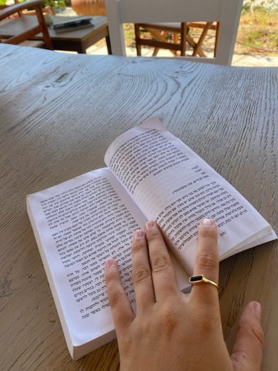An overhead photograph captures an open book resting on a light-colored, weathered wood-grain table. A human hand, positioned with fingers splayed, rests on the lower right page of the book. The hand wears a gold-toned ring with a black, rectangular, horizontally set stone on the ring finger. The book's pages display text in Hebrew script, with the page number "168" visible at the bottom right corner of the right-hand page. The background is out of focus, revealing elements consistent with an outdoor or semi-outdoor setting, including parts of a white wooden chair, a natural wood-colored chair, green foliage, and a terracotta pot, indicating a well-lit environment. The scene is located in Πλατύς Γιαλός, Greece.