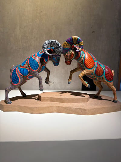 Two brightly painted wooden ram sculptures are mounted on a neutral wood panel affixed to a wall. The lower ram, decorated with blue and orange patterns, faces upward. Above it, the second ram, adorned with orange and turquoise designs, faces downward. The ram's horns are intricately detailed with multiple colors and patterns. The wall behind the panel is divided into two sections, one a smooth white, and the other a textured grey.