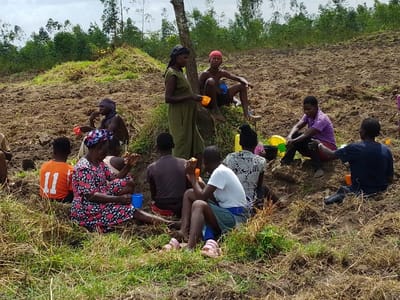 A group of people sit and relax in a field after work. The area is a mixture of sparse grass and tilled dirt with trees in the background. Most of the individuals have cups in their hands, implying they are enjoying a drink or snack. Some are conversing, while others are resting quietly. They are a mix of men and women, dressed in casual clothing. A clear day is depicted with a cloudy sky.
