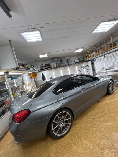 A grey BMW coupe is parked in a garage. The car is facing right, with its rear on the left side of the frame. The interior of the garage is visible, with shelves stocked with various items, including bottles and cleaning supplies. There are fluorescent lights on the ceiling. The floor is a speckled yellow material. The garage door is visible in the background on the right. No people are present in the image.
