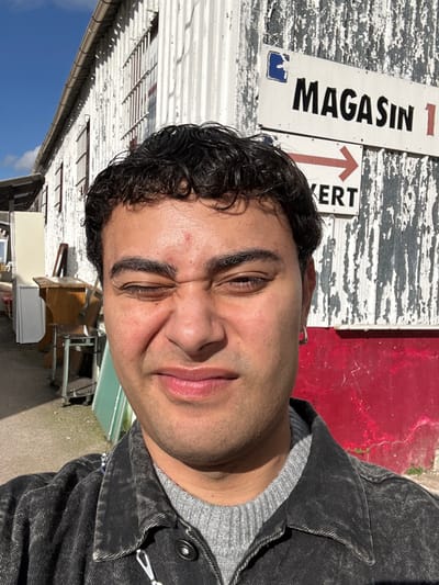 A male individual is depicted in a close-up selfie-style photograph, positioned outdoors under direct sunlight in Neuilly-Plaisance, France. The subject, appearing to be an adult, possesses dark, curly hair and a fair complexion with a minor blemish on the forehead. He is wearing a grey ribbed sweater beneath a faded dark grey or black denim jacket. A silver chain is visible on his left side, and a silver hoop earring is present in his right ear. His facial expression involves a pronounced squint of the left eye (viewer's right), a partially closed right eye (viewer's left), and a crinkled nose and forehead, indicating a wince or strong facial contortion.
The background features the exterior of a utilitarian building, constructed with corrugated metal or siding. The upper portion of the wall is painted white, extensively worn and peeling to reveal underlying grey material. The lower section of the wall to the right is painted a vivid red, also showing significant weathering. Multiple window openings with vertical bars are visible on the white wall. Two rectangular signs are affixed to the white siding: an upper sign reads "MAGASIN 1" in dark and red lettering, accompanied by a blue graphic on the left; a lower sign displays a prominent red arrow pointing to the right, with partially visible text "KERT" beneath it.
To the left of the subject, an accumulation of discarded or stored items is visible, including a tall white cabinet, a wooden table, and various other objects, suggesting a storage area, reclamation yard, or second-hand establishment. The sky above is clear blue with some scattered white clouds.