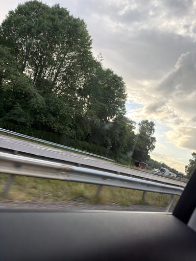 The image depicts a view from inside a vehicle looking out towards a highway. On the left side of the highway, there is a dense line of tall, green trees. The highway itself has two lanes visible, separated by white lines, and is bordered by a silver metal guardrail in the foreground and middle ground. Beyond the highway, the terrain is covered with green grass and some dry patches. In the distance, on the right side of the highway, several vehicles are visible, including trucks and cars. The sky is filled with clouds, with patches of sunlight breaking through, suggesting either early morning or late afternoon. A small red building is also visible near the highway. The scene is located on a highway in or near Frillesås, Sweden.