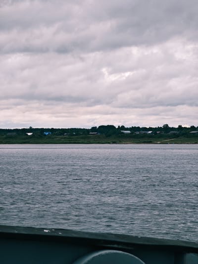 The image shows a wide, choppy river with a village on the far bank. The sky is overcast with heavy, gray clouds. The village consists of several wooden houses with various colored roofs, situated on a grassy embankment lined with trees. Some boats are visible along the shoreline of the village. In the foreground, the dark, blurry edge of what appears to be a vessel is visible, suggesting the photo was taken from a boat on the river. The overall scene is calm and somewhat somber, typical of a cloudy day in a rural setting. The location is a village on the Yenisei River in Zotino, Krasnoyarsk Krai, Russia.