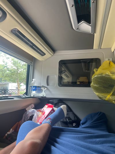 The image shows the interior of a vehicle, likely a bus or van, with a person's legs visible in the foreground, covered by a blue blanket. The person is wearing blue socks with a pattern. Adjacent to the legs is a red and white box of "Choco Pie". To the right, a yellow plastic bag filled with dark items is hanging. A small black screen is mounted on the wall of the vehicle, displaying a blurry image. On the left, a window looks out onto trees and some buildings, suggesting a semi-urban or rural setting. The interior of the vehicle features cream-colored upholstery and air vents. Based on the context provided by the prompt, this scene is located in Pờ Y, Vietnam.