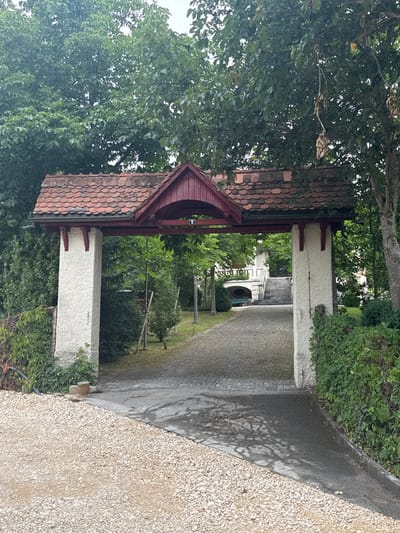 This image captures an entranceway to a property, likely in Tavannes, Switzerland. The entrance is an archway supported by two thick, white, stuccoed pillars. The archway itself is topped with a red-tiled roof, a common architectural style in Swiss homes. The roof has a dark red, wooden trim and is supported by decorative wooden brackets.
Beyond the archway, a cobblestone driveway leads up to a white house with a grand staircase. The house appears to be surrounded by lush green trees and manicured bushes. The overall impression is one of a well-maintained, affluent property. The scene is serene and tranquil, suggesting a peaceful rural setting. The sky is overcast, indicating a possibly mild or rainy day. The image appears to have been taken during daylight hours.