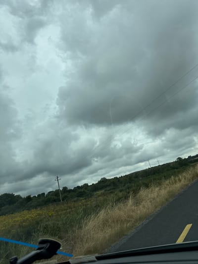 The image captures a view from a vehicle traveling on a road in Scartnamore, Ireland. The sky is filled with heavy, dark clouds, suggesting an overcast or approaching storm. A faint, arc-shaped rainbow is visible through the clouds, partially obscured. Power lines stretch across the sky, supported by wooden poles. The roadside is lined with dry, tall grass and scattered yellow wildflowers. Beyond the immediate roadside, a green, undulating landscape with trees and bushes rises towards the horizon. The road itself is paved and dark, with a distinct yellow line marking the edge. The lower part of the image shows the dashboard of the vehicle, including a black electronic device mount with a blue sticker.