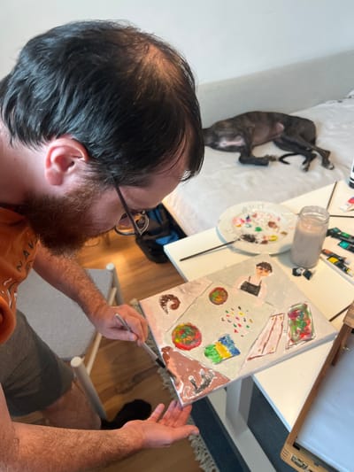 A man with dark hair and a beard, wearing glasses and an orange t-shirt, is painting on a canvas. He is sitting at a white table, holding a paintbrush in his right hand and a palette in his left. The painting on the canvas depicts a person wearing an apron, surrounded by colorful shapes and jars.

On the table, there is a white plate with various paint colors, a jar of liquid, tubes of paint, and other painting supplies. In the background, a dog is sleeping on a bed. The floor is made of wood. The environment appears to be an indoor setting, possibly a home or studio. There is no specific indication of the location being in Hämeenlinna, Finland.