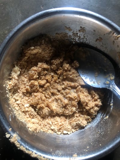 The image shows a close-up, overhead shot of a stainless steel bowl containing a mixture of beige and light brown, irregularly shaped flakes or granules, possibly food ingredients. A silver spoon rests on top of the mixture. The bowl is placed on what appears to be a weathered, possibly concrete or stone surface. There are no people visible in the frame. The image does not provide any information to determine a location in Idima, Nigeria.