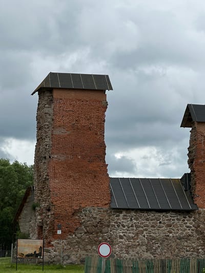 The image shows the exterior of a partially ruined brick and stone structure, likely a historical landmark, under a cloudy sky. The architecture features weathered red brickwork interspersed with rough-hewn stones, indicating age and decay. Several sections of the walls are missing, revealing the inner structure and the sky beyond. Each remaining section of the wall is topped with a sloped, dark-colored roof, possibly made of metal or shingles, with visible support beams. In the foreground, a green wooden fence runs horizontally across the frame. To the left, a signpost with an informational display panel is situated on a grassy area. A circular white sign with a red border is also visible to the right of the fence. The environment suggests an outdoor setting, possibly a historical site or park, given the presence of the informational sign and the protective fence. The overall impression is one of historical preservation and enduring architectural presence despite the passage of time.