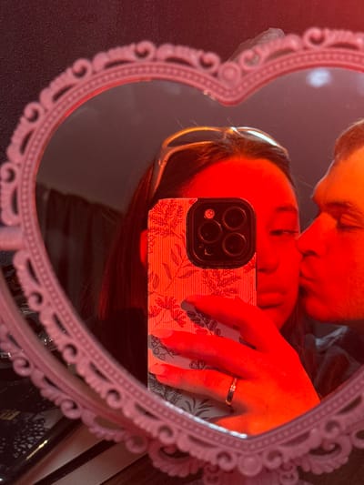 A mirror selfie captures two individuals reflected in an ornate, light-colored, heart-shaped frame. The entire scene is illuminated with a dominant red light. A person with dark hair, wearing sunglasses on their head, holds a smartphone with a floral-patterned case and a triple camera array, obscuring the lower half of their face. Their right hand, adorned with a gold-colored ring on the ring finger, holds the device. A second individual is partially visible on the right, kissing the first person's cheek. The background surrounding the mirror is dark and indistinct. The specific location within Prikubanskiy, Russia, is not identifiable from the visual evidence provided.