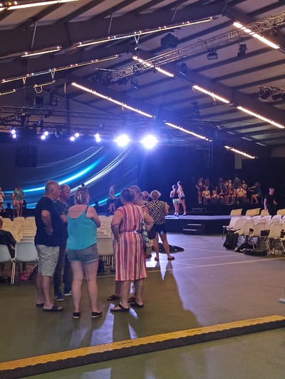 This media file captures an indoor event, likely a performance or gathering, in Warffum, Netherlands. The scene is dominated by theatrical lighting, with rows of bright, long fluorescent lights illuminating the space from above. A stage area is visible in the background, featuring what appears to be a large screen with a graphic of swirling blue light.
In the foreground and mid-ground, several groups of people are standing. They appear to be attendees, with some facing the stage and others interacting amongst themselves. The attire of the individuals suggests a casual event, with t-shirts, shorts, and dresses being common. One group in the foreground consists of three people, two women and one man, all facing away from the camera and towards the stage. A woman in a blue tank top and grey shorts stands beside a man in a dark t-shirt and grey shorts. To their left, another woman in a striped dress is visible.
Behind these individuals, more people are gathered, some seated in white chairs that are arranged in rows, and others standing. The lighting casts long shadows, indicating the artificial nature of the illumination rather than natural daylight. The overall atmosphere seems to be one of anticipation or engagement with an event unfolding on the stage. The presence of stage lighting and a backdrop screen strongly suggests a planned performance, concert, or presentation. No specific text is clearly legible in the image.