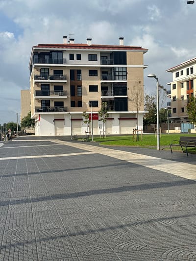 The image depicts a modern urban scene in Pineda de Mar, Spain, featuring a multi-story building alongside a public plaza and green space under a partly cloudy sky.

The central subject is a five-story residential building with a beige facade, white trim, and a terracotta-tiled hip roof with multiple dark chimney stacks. The upper four floors feature balconies with dark-framed glass railings and large windows, some with dark roller shutters. The ground floor consists of several commercial units, all secured by closed light-colored roller shutters, indicating they are currently shut. A red sign partially visible on one unit displays "[U Tic]".

In the foreground, a patterned paved plaza extends from the bottom of the frame, constructed from dark grey interlocking pavers with subtle circular and grid patterns. Lighter grey plain paving stones form pathways within this dark grey area. To the right of the plaza is a well-maintained grassy area containing several young trees supported by wooden stakes. Two dark-colored public benches are positioned within this green space: one on the right side of the frame, and another closer to the building on the left. Multiple contemporary streetlights with curved tops are visible throughout the scene.

In the far left background, two individuals are seen walking away from the viewer on a paved path adjacent to a road, partially obscured by a tree. Another similar multi-story building, also with a terracotta roof, is partially visible in the right background. The sky above is overcast with patches of blue visible. No specific actions or interactions among the visible people are discernible.