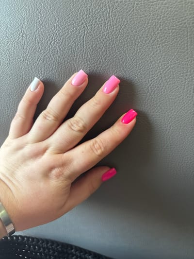 A person's hand, with long, square-shaped artificial nails painted in various bright colors, is resting on a textured gray surface, possibly a couch or car seat. The nails are painted in white, light pink, bright pink, and neon pink. The hand is positioned with the palm facing down, and the fingers are slightly spread. The background is out of focus, but appears to be a continuation of the gray textured surface. There is a black woven material visible at the bottom edge of the image, and a silver bracelet on the person's wrist. The image appears to be taken indoors in natural light. There is no specific information about the location in Мастоцкі сельскі Савет, Belarus.