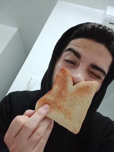 This is an informal, close-up portrait of a person in a minimalist indoor setting, likely a home in Fgura, Malta. The subject is a young adult with dark hair, wearing a black hooded sweatshirt. They are holding a single slice of lightly toasted white bread in front of their face, playfully covering their nose and mouth. Their left eye is open and winking, conveying a humorous or lighthearted mood, while their right eye is largely obscured by the toast. The person's left hand, holding the toast, is visible in the foreground.
The background consists of clean, light-colored walls, primarily white and light grey, forming distinct geometric lines and corners. On the white wall behind the subject's right shoulder, a white rectangular device is mounted. This device features a small red dot or indicator light and several faint grey lines or markings, suggestive of an electrical outlet, thermostat, or sensor, but no legible text is visible. The lighting is bright and even, indicating an indoor daytime environment.