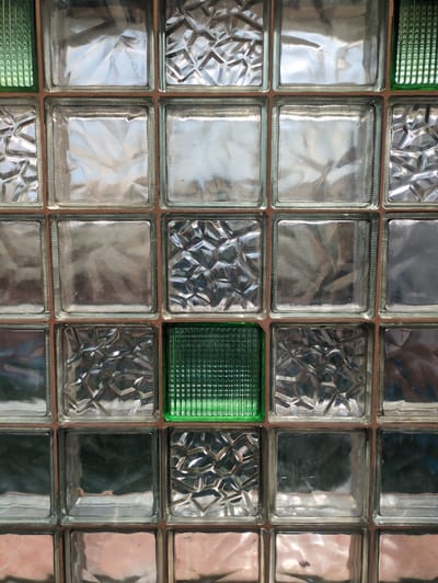 This media file presents a close-up, overhead view of a wall constructed from glass blocks. The blocks are arranged in a grid pattern. Most of the glass blocks are clear and reflect light, showing various textured surfaces and creating a mosaic-like appearance. Some blocks have a faceted, geometric pattern, while others appear smooth or lightly textured, causing the reflections to be somewhat distorted. Two of the blocks are a vibrant green color and have a ribbed texture, providing a contrast to the predominantly clear blocks. The blocks are framed by a reddish-brown material, likely grout or mortar, holding them together. The lighting suggests it is daytime, with the bright reflections indicating a light source directly above or in front of the wall. The overall scene is an abstract composition of light, texture, and color within the structured grid of glass blocks. There is no visible text, people, or discernible activity or event taking place. The setting appears to be an interior wall, possibly in a building in Velikovo, Russia, given the location context, but this cannot be definitively determined from the image alone.
