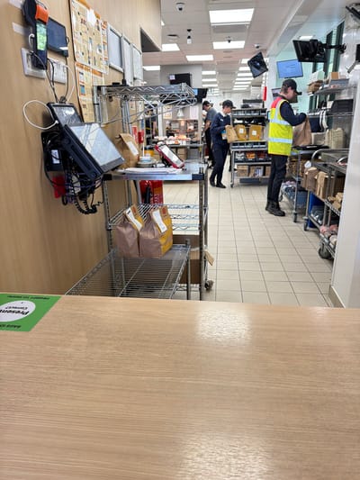 The image depicts an interior view of what appears to be a commercial kitchen or storage area in a business located in Pity Me, United Kingdom. Two workers are visible in the background. One is wearing a high-visibility vest and appears to be handling items on a shelving unit. Another worker is dressed in darker clothing and also seems to be interacting with stored items. Shelving units filled with various items line the walls. In the foreground, there is a countertop with technological devices, including touch screen monitors and a payment terminal. A document or schematic is laid out on the counter. Bags are staged for delivery behind the workstation. A section of a wooden wall with a green sticker is prominent in the foreground.