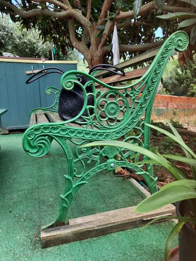 A green, ornate, cast-iron bench with wooden slats sits on a green, artificial turf surface. A black handbag rests on the bench. In the background, a green shed is visible, as well as trees, foliage, and a brick wall. The scene is set in a garden in Pesaro, Italy.