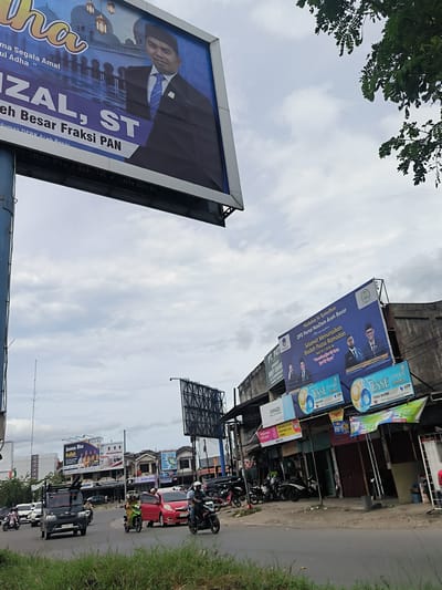 This media file captures a bustling street scene in Lambaro, Indonesia, under an overcast sky. The image shows a road with a grassy median, populated by various vehicles including a black van (license plate BL 8396 LN), a red car, and several motorcycles with riders, indicating active traffic.
Two prominent political billboards dominate the upper half of the image. The large blue billboard on the left features a man identified as "RIZAL, ST" from "Fraksi PAN", with text related to "Idul Adha" and "Humas DPRK Aceh Besar". Another large blue billboard on the right, affixed to a commercial building, displays multiple men and carries the message "Marhaban Ya Ramadhan" and "Selamat Menunaikan Ibadah Puasa Ramadan 1447 H / 2026 M" from "DPD Partai NasDem Aceh Besar".
Below this second billboard, a row of commercial establishments lines the street, featuring various signs. These include "JUMADI", "MEDIA CELL", "MONEY CHANGE", and "ACEH POS". Several advertisements for "ESSE CHANGE DOUBLE" cigarettes are also visible. In the distance, another billboard or sign advertises "Semua Bisa Kuliah".
The sky is cloudy, suggesting a daytime scene without direct sunlight. Tree foliage is visible in the upper right corner, adding a natural element to the urban landscape. The overall scene depicts a busy commercial and transportation hub.