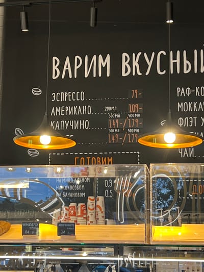 This is a medium shot photograph of a coffee shop's menu board and display case. The menu board is black with white lettering listing coffee drinks like "ЭСПРЕССО," "АМЕРИКАНО," and "КАПУЧИНО," along with prices and sizes. To the right, other coffee drinks like "РАФ-КО" and "МОККА" are listed. Below the main menu, "ГОТОВИМ" is written in orange, indicating food preparation. In the display case below, there are labels for "ЧИАБАТТА" and "МИНИ ЧИАБАТТА" with prices. There are also stacks of coffee cups and reflections of trees and the interior of the shop in the glass. The overall environment suggests a casual, modern cafe. There are no people clearly visible in the frame. The image was taken in Косой Карьер Снт, Russia.