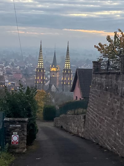 An elevated view captures a misty twilight or early morning scene in Obernai, France. In the far background, under an overcast sky with a faint horizon band of pale orange light, three illuminated church spires dominate the skyline. Two tall, slender spires, belonging to the Church of Saints Peter and Paul, stand prominently with vertical rows of yellowish lights, flanking a slightly shorter, broader central spire also lit. Below these, the hazy expanse of Obernai's rooftops and buildings is visible, dotted with faint artificial lights. Beyond the town, a flat, misty landscape extends to the horizon.

In the foreground, a dark, narrow paved path, identified as a "CHEMIN PRIVE" (Private Road) by a red and white circular sign affixed to a stone pillar, curves uphill from the lower center. To the left of the path, dense green foliage, including hedges and trees, lines the ascending terrain. The stone pillar in the lower left corner also displays the number "9" and likely functions as a mailbox support. To the right of the path, a substantial stone wall, constructed from rough-hewn blocks, runs diagonally, ascending alongside the path. Above this wall, a dark-roofed house with pink-colored walls is partially visible, behind which a section of metal railing and a small tree with autumn-colored foliage are present. No people, animals, or vehicles are observed within the frame. A thin wire traverses the upper left quadrant of the sky.