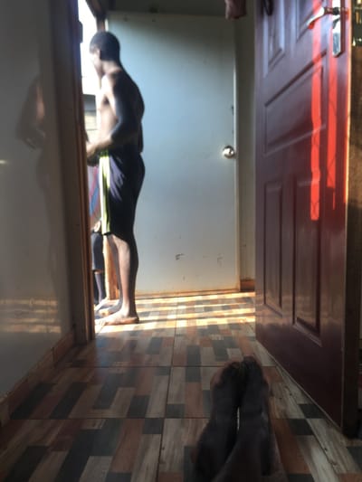 An indoor scene, contextually identified as Riana ward, Kenya. The perspective is low, revealing the crossed, dark-skinned feet of an individual in the immediate foreground. To the right, a dark red paneled door is ajar, casting distinct vertical shadows across the floor. The floor is tiled with a patterned surface featuring rectangular shapes in tones of brown, grey, and black. Through an open, light-colored doorway on the left, a shirtless dark-skinned male is visible, standing with his back to the viewer, wearing dark shorts with a light vertical stripe on the side. A faint reflection of his upper body is present on a reflective surface to the left of the doorway. Partially obscured behind him, the lower legs of a second individual are discernible. Beyond the figures, a plain light-colored wall with a metallic door handle is visible. Strong, directional sunlight illuminates the area through the open doorway, creating sharp patterns of light and shadow on the tiled floor.