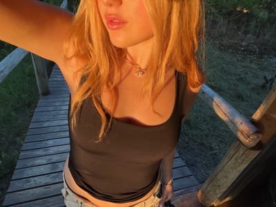 The image shows a woman with blonde hair hanging upside down, likely taking a selfie on a wooden deck or platform. She is wearing a black tank top and a bracelet on her left wrist. She has a gold necklace with a small round pendant. The background is out of focus, but there is evidence of grass and wooden supports of the structure. The lighting suggests it is either sunrise or sunset. The specific location in Khwai, Botswana cannot be confirmed from the image.