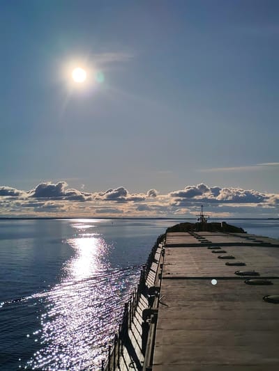 A long, flat deck extends to the horizon, with a railing on the left and metal plates covering the deck. The sea sparkles in the sunlight on the left, and puffy clouds are on the horizon. The sun is high in the sky, casting a glare. The location is Vichelovo, Russia, based on the mention of the city.