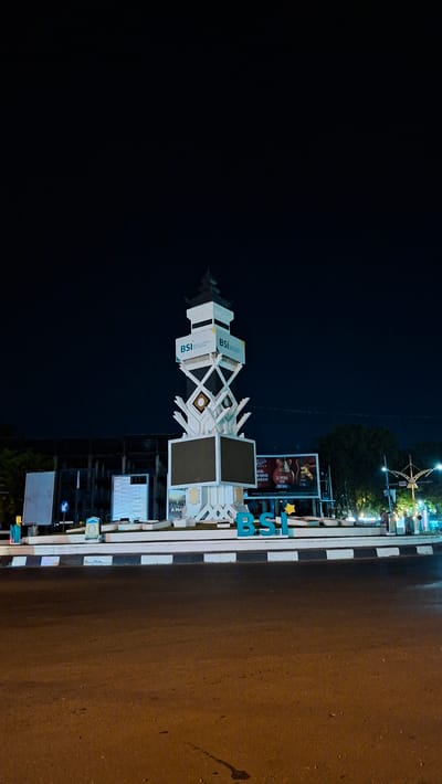 The image shows a monument at night in Lamprehlam Teungoh, Indonesia. The monument is a tall, white, multi-tiered structure with geometric patterns and several large screens. The words "BSI" are prominently displayed in blue letters at the base of the monument, and also on the sides of the upper tiers. There are several streetlights and some trees in the background, as well as some buildings and billboards. A few people are visible walking around the area. The road in the foreground is asphalt and appears wet, reflecting the lights from the surroundings. The sky is dark and clear, indicating nighttime.