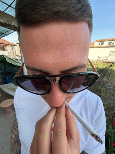 A close-up, slightly tilted shot features a man holding a lit cigarette between his lips and fingers. He is wearing black Carrera sunglasses with gold accents. He has dark hair and light skin. The man is wearing a white t-shirt. His tattooed arm is visible. The background includes foliage, tulips, a building, and a fence. The location may be Damianitsa, Bulgaria.