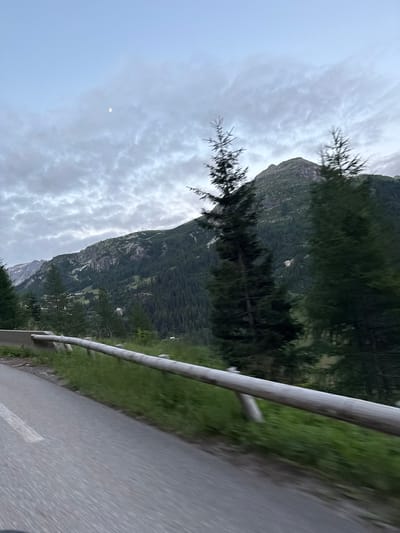 The image depicts a mountain landscape with a road in the foreground. The road is paved and has a white line marking the lane. A wooden guardrail runs alongside the road, separating it from a grassy embankment. In the background, a densely forested mountain rises, with a lighter-colored peak visible in the distance. The sky is overcast with some patchy clouds, and a small, faint moon is visible. The overall impression is of a scenic mountain view, possibly taken from a moving vehicle due to the motion blur. There are no people or specific structures visible that would indicate a city location. The query's request to place this image in Tignes, France, cannot be verified from the visual information provided.