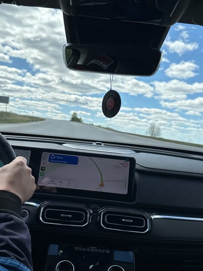 The image shows a view from inside a car, with the dashboard and parts of the steering wheel visible. The navigation system displays a map with a route indicating "1212 GM, Pulyanovskata" as the next direction, and the destination is "Doma 78 Km" with an estimated time of arrival at 11:00. In the rearview mirror, the reflections of a cloud-filled sky are visible, and a circular air freshener hangs from the mirror. The driver's hand is visible on the steering wheel.