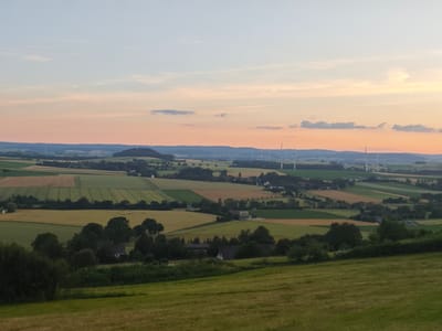 The image depicts a panoramic view of a rural landscape during sunset in Schieder-Schwalenberg, Germany. The sky is a soft gradient of blue and orange, with wispy clouds. In the distance, rolling hills are dotted with wind turbines. The foreground is dominated by a lush green meadow, with the middle ground showing a patchwork of agricultural fields, trees, and some scattered buildings, likely farms. A small building with a dark roof and white walls is visible amongst the trees in the mid-ground. The overall impression is one of tranquility and natural beauty.
