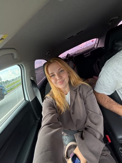 The image shows a young woman with blonde hair and fair skin, wearing a brown blazer over a white top, seated in the passenger seat of a car. She is smiling at the camera, which is held by an unseen person. Outside the car window, a highway with a blue tractor is visible, suggesting the car is in motion. The interior of the car features black upholstery and a grey headliner. A portion of another person's arm, possibly a male, wearing a grey t-shirt, is visible on the right side of the frame. The scene appears to be taking place in or around Kuznechikha, Russia, based on the provided location.