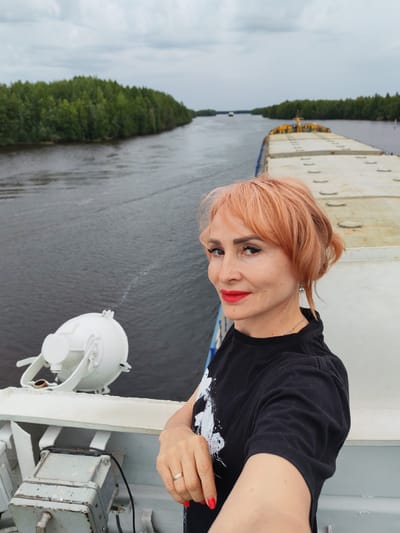 A woman with short, coral-colored hair and red lipstick is taking a selfie on the deck of a ship. She is wearing a black t-shirt with a white graphic design and a silver necklace. She is also wearing a ring on her left hand, which is visible as she holds the camera.
The woman is positioned on the right side of the frame, with the open water of a river to her left and the deck of a cargo ship to her right. The river is flanked by dense green trees on both sides, indicating a natural, wooded environment. In the distance, another cargo ship can be seen on the river. The sky is overcast, suggesting a cloudy day.
The setting appears to be a waterway, possibly a canal or river, in a region with abundant forests. The presence of large cargo ships implies commercial or industrial activity related to water transport. The overall mood is casual and relaxed, with the woman appearing to enjoy her time on the ship. No visible text is present in the image.