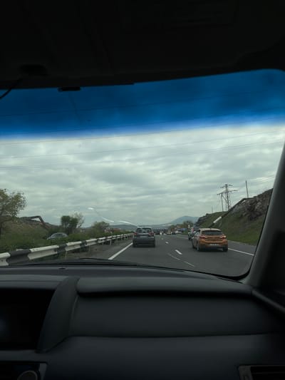 Captured from inside a vehicle, the image presents a view of a road scene in Arzni, Armenia. The road shows light traffic consisting of cars, with some appearing to be stopped in the roadway. The sky is overcast, creating a dull background over the scene. Infrastructure along the roadside includes utility poles.