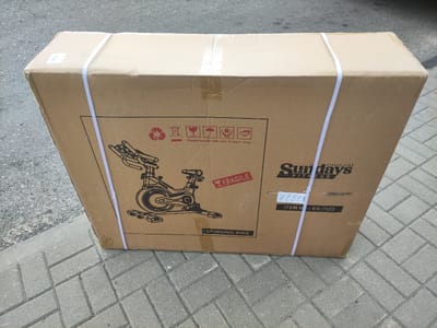 A large cardboard box containing a "SPINNING BIKE" from "Sundays FITNESS" is resting on a paved walkway. The box is secured with white plastic strapping. Various handling symbols, including "Please handle with care & don't drop" and a "FRAGILE" sticker, are printed on the box. The box has some wear and tear, with tape visible on the seams. The background appears to be an outdoor setting with asphalt and brick paving. The image is taken from a slightly elevated angle, looking down at the box. The item number "ES-7022" is clearly visible. There are no people visible in the image. The lighting suggests it might be daytime.